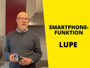 Mann mit grauem Pullover steht in Küche mit Smartphone, rechts Text auf gelbem Hintergrund: "Smartphonefunktion Lupe"