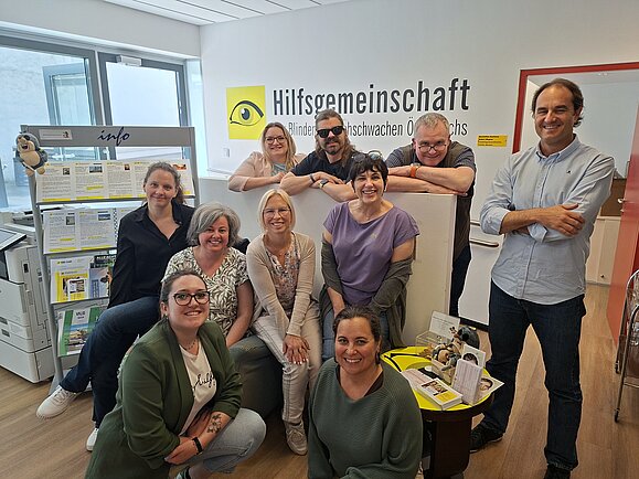 Mehrere Personen stehen, hocken bzw. sitzen und lächeln in den Räumlichkeiten der Hilfsgemeinschaft in die Kamera.