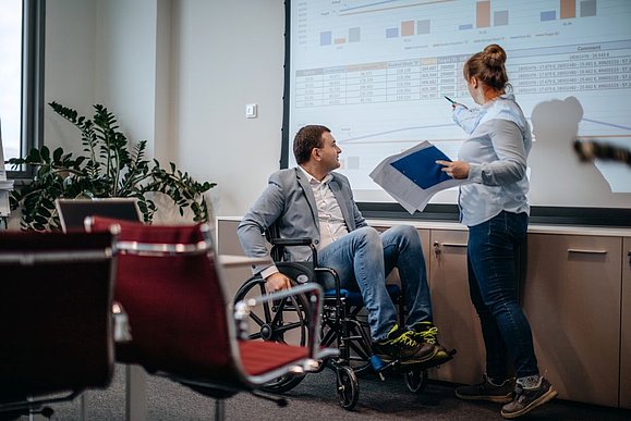 Mann mit Rollstuhl und stehende Frau im Büro schauen sich Zahlen auf einer Leinwand an