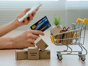 kleiner Einkaufswagen mit Lieferpaketen gefüllt, im Hintergrund eine Hand die Smartphone und Kreditkarte hält