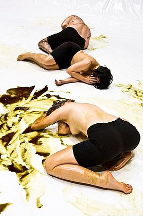 drei Menschen liegen am Boden mit schwarzer kurzer Hose und nacktem Oberkörper, die in goldene Streifen tauchen