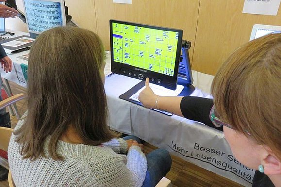 Junge Frau sitzt vor einem Bildschirmlesegerät und lässt sich von einer Beraterin die auf den Bildschirm zeigt dieses erklären.