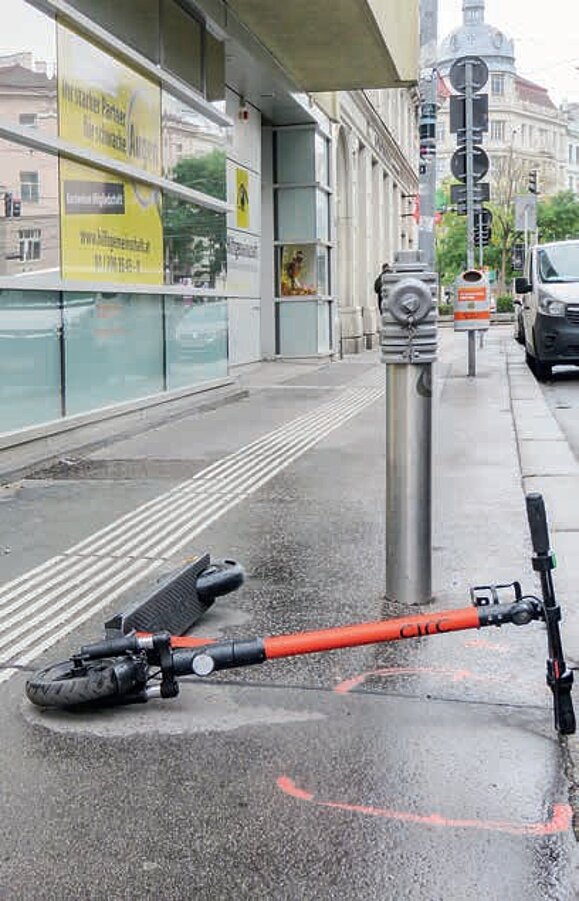 Ein oranger E-Scooter liegt auf einem Gehsteig direkt neben dem Blindenleitsystem.