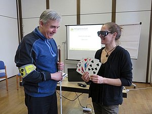 Petra Wrba hält große Tarockkarten in den Händen, sie trägt dabei eine Simulationsbrille. Harald Rother steht mit Taststock und Armbinde daneben.