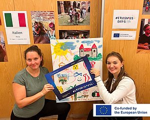 Eröffnung der Fotoausstellung Zwei junge Frauen halten einen Bilderrahmen, im Hintergrund sieht man Fotos