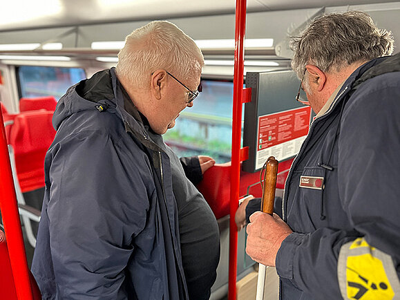 OEBB_Cityjet_Elmar_Fuerst_2025 Zwei Männer stehen in einem modernen Zug vor einem Knopf. Beide tragen Jacken, einer hält einen Taststock und trägt eine gelbe Armbinde. Er drückt den Knopf, während ihn beide betrachten.
