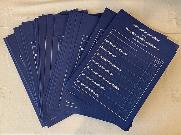 Blaue Wahlschablonen mit weißer Schrift in A4 liegen ausgebreitet auf dem Tisch. Ein ausgestanztes Kasterl je Kandidat