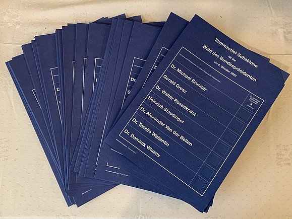 Blaue Wahlschablonen mit weißer Schrift in A4 liegen ausgebreitet auf dem Tisch. Ein ausgestanztes Kasterl je Kandidat