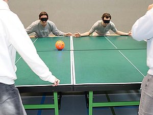 Zwei Menschen mit Augenbinden stehen hinter zwei zusammengeschobenen Tischtennistischen. Sie beugen sich mit ausgebreiteten Armen zum Tisch hinunter, um zu verhindern, dass ein Ball vom Tisch rollt.
