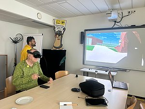 Virtuelle Realität für Mobilitätstraining sehbehinderter Menschen Ein Mann hat eine virtuelle Brille oben, man sieht einen Bilschirm. Neben dem Bildschirm steht noch ein Mann.