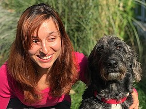 Frau mit langen braunen Haaren und pinkem Shirt lächelt in die Kamera und hat ihren Arm um einen schwarzen Hund mit krausen Haaren