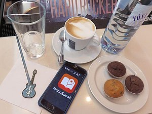 Kaffee, Muffins und Mineralwasser stehen auf einem Tisch und die App Menu Speak ist auf einem Smartphone aktiviert