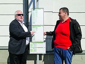 Zwei Männer mit schwarzen Jacken zeigen auf ausgehängten Busplan