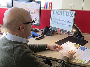 Mann sitzt mit vergrößerndem Lesegerät am Arbeitsplatz