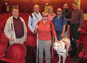 Gruppe von Menschen in rotem Theatersaal teils mit Brillen, Langstock und vorne Frau mit Blindenführhund