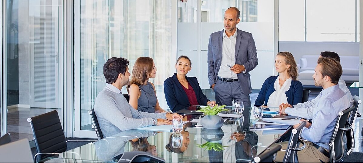 Ein Mann spricht vor mehreren Menschen, die rund um einen Tisch sitzen.