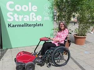 Maria Grundner sitzt im Rollstuhl vor der grünen Plakatwand der "Coolen Straße" am Karmeliterplatz.