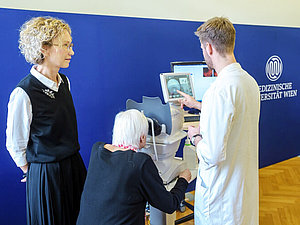 Eine ältere Frau schaut in ein Gerät, dass von einem jungen Herrn bedient wird. Daneben steht eine blonde Frau. 