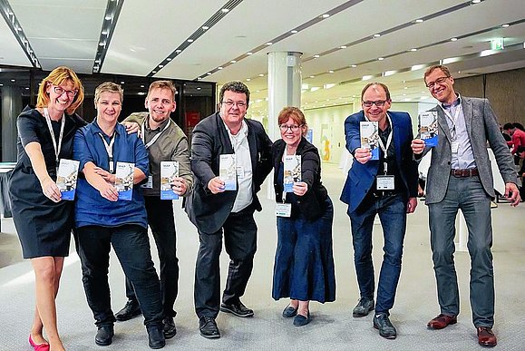 7 Menschen in Businesskleidung und mit Umhängeschild strecken Flyer mit Aufschrift "IAAP" in die Kamera.